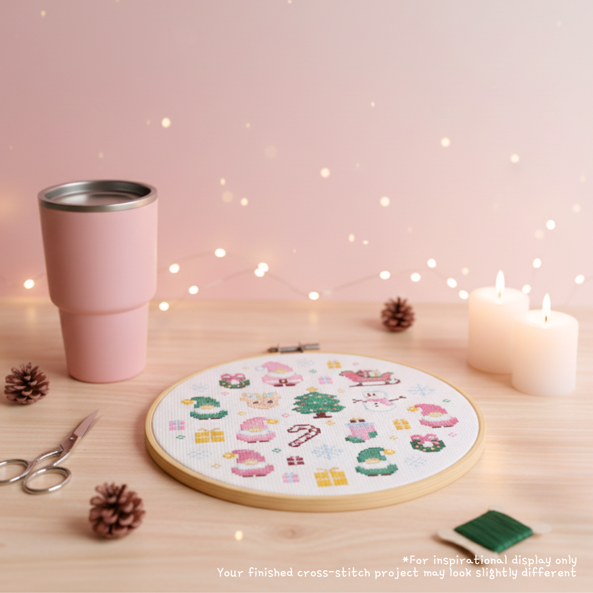 Finished pink Santa cross stitch in a hoop on a wooden table with candles, scissors and a pink cup